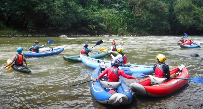 A group of people wearing safety gear paddle individual watercraft on a river. 